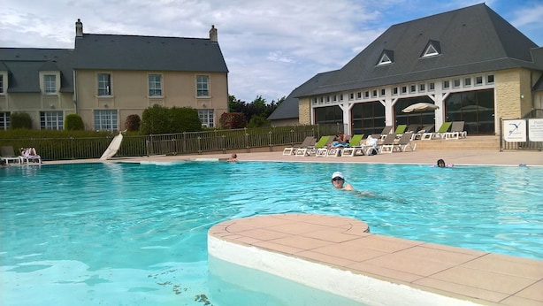Piscina com cadeiras de descanso e um edifício com telhado inclinado ao fundo.