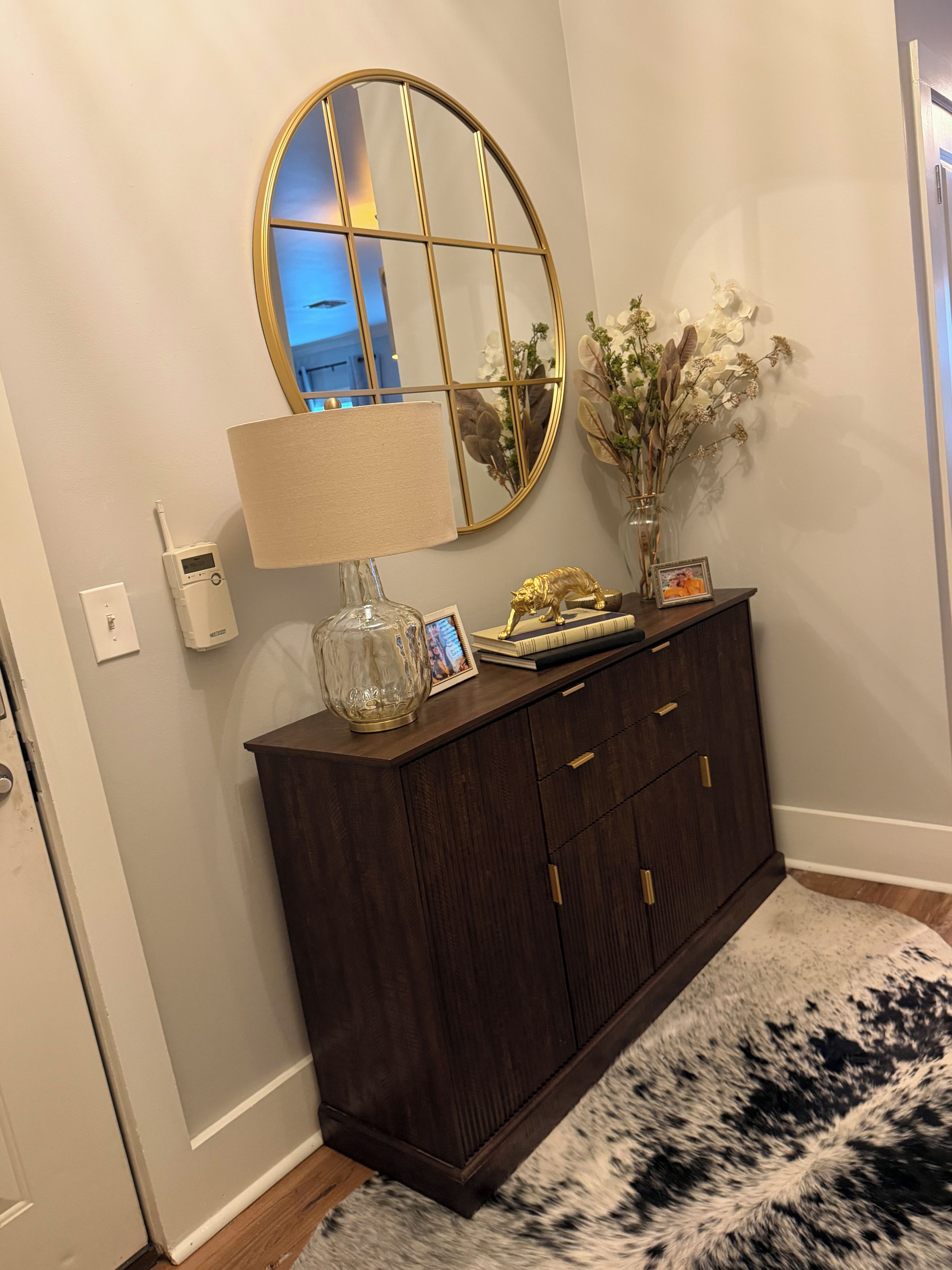 A dark wood cabinet with a round mirror, a lamp, a small statue, and a vase with dried flowers.
