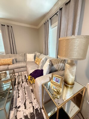 A living room with a sectional sofa, a glass coffee table, a patterned rug, a gold lamp, and a framed photo.