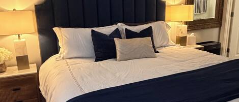 Un lit bien fait, avec une tête de lit sombre, du linge de lit blanc et plusieurs oreillers. On aperçoit une table de chevet avec une lampe et un vase de fleurs. Un miroir est visible sur le mur, à droite.