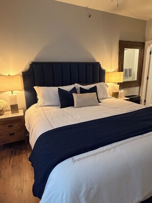 A neatly made bed with a dark headboard, white bedding, and several pillows. There is a bedside table with a lamp and a vase of flowers. A mirror is visible on the wall to the right.