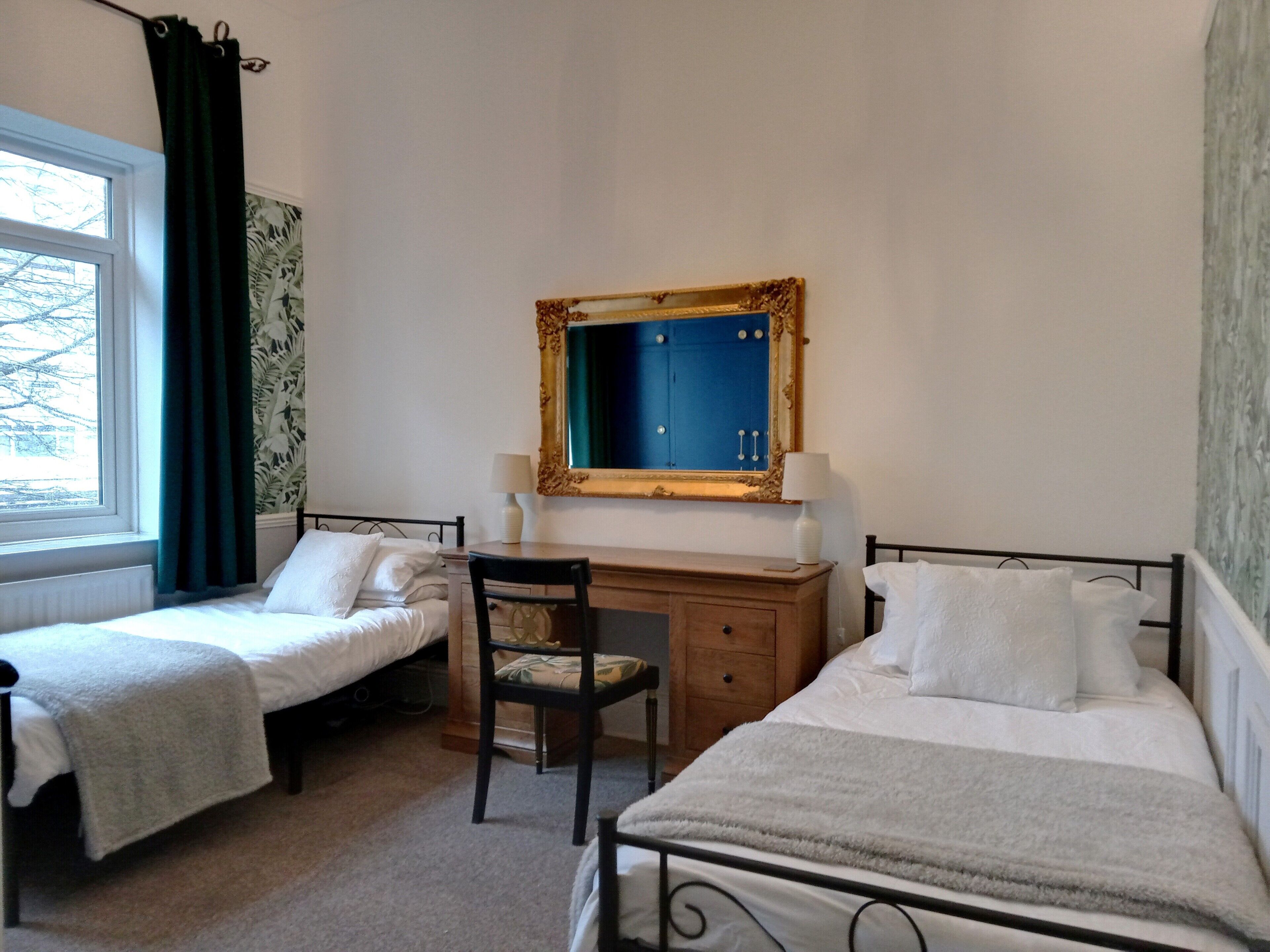 A room with two single beds, a desk with a chair, a mirror, and a window with curtains.
