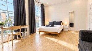 A modern bedroom with a wooden bed, a desk, and a chair. There is a window with curtains, a bedside table with a lamp, and a view of buildings outside. - Spacious 3BR Walk to Camden, Euston & King’s Cross (Greater London)