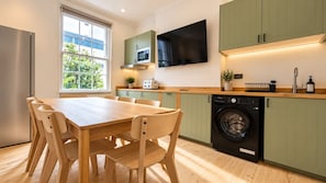 A modern kitchen with a wooden table and chairs, a washing machine, and a flat-screen TV mounted above the counter. - Spacious 3BR Walk to Camden, Euston & King’s Cross (Greater London)