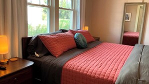 Quarto com cama, mesas de cabeceira, cômoda, espelho e janela com cortinas.