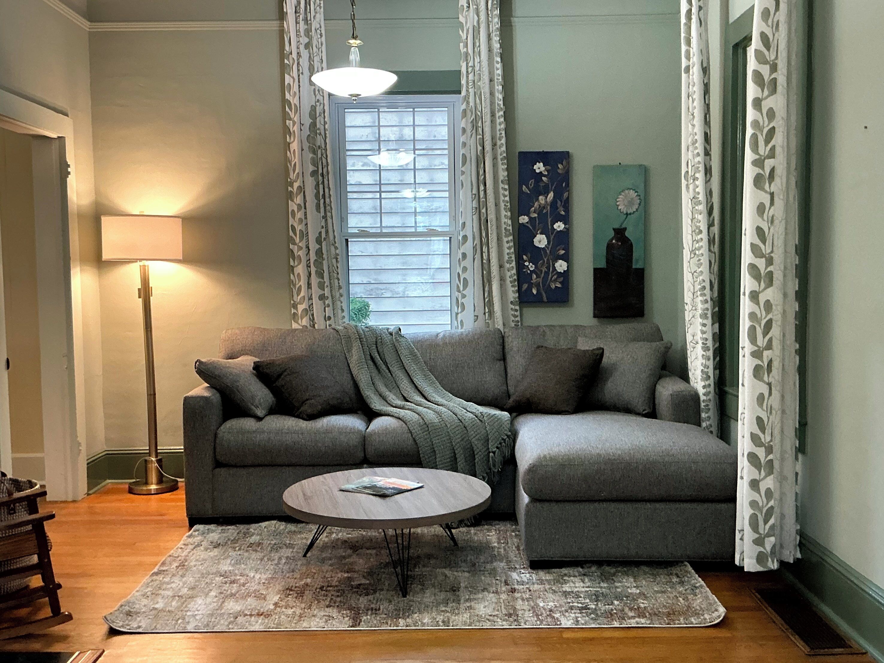 Un salon avec un canapé d’angle gris, une table basse ronde, un lampadaire et une fenêtre avec des rideaux.