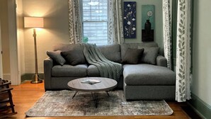 Sala de estar com sofá modular cinza, mesa de centro redonda, luminária de chão e janela com cortinas.