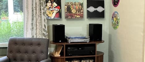 A room with a grey armchair, a wooden cabinet with vinyl records, and a window with curtains.