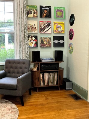 Ein Zimmer mit grauem Sessel, einem Holzschrank mit Schallplatten und einem Fenster mit Vorhängen.