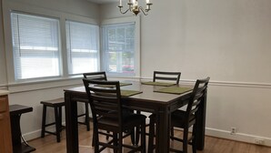Uma área de refeições com mesa e cadeiras de madeira, um lustre e janelas com persianas.