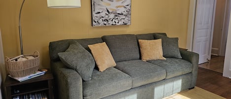 Un sofá gris con dos cojines, una lámpara de pie, una estantería llena de libros, una obra de arte enmarcada y un piso de madera.