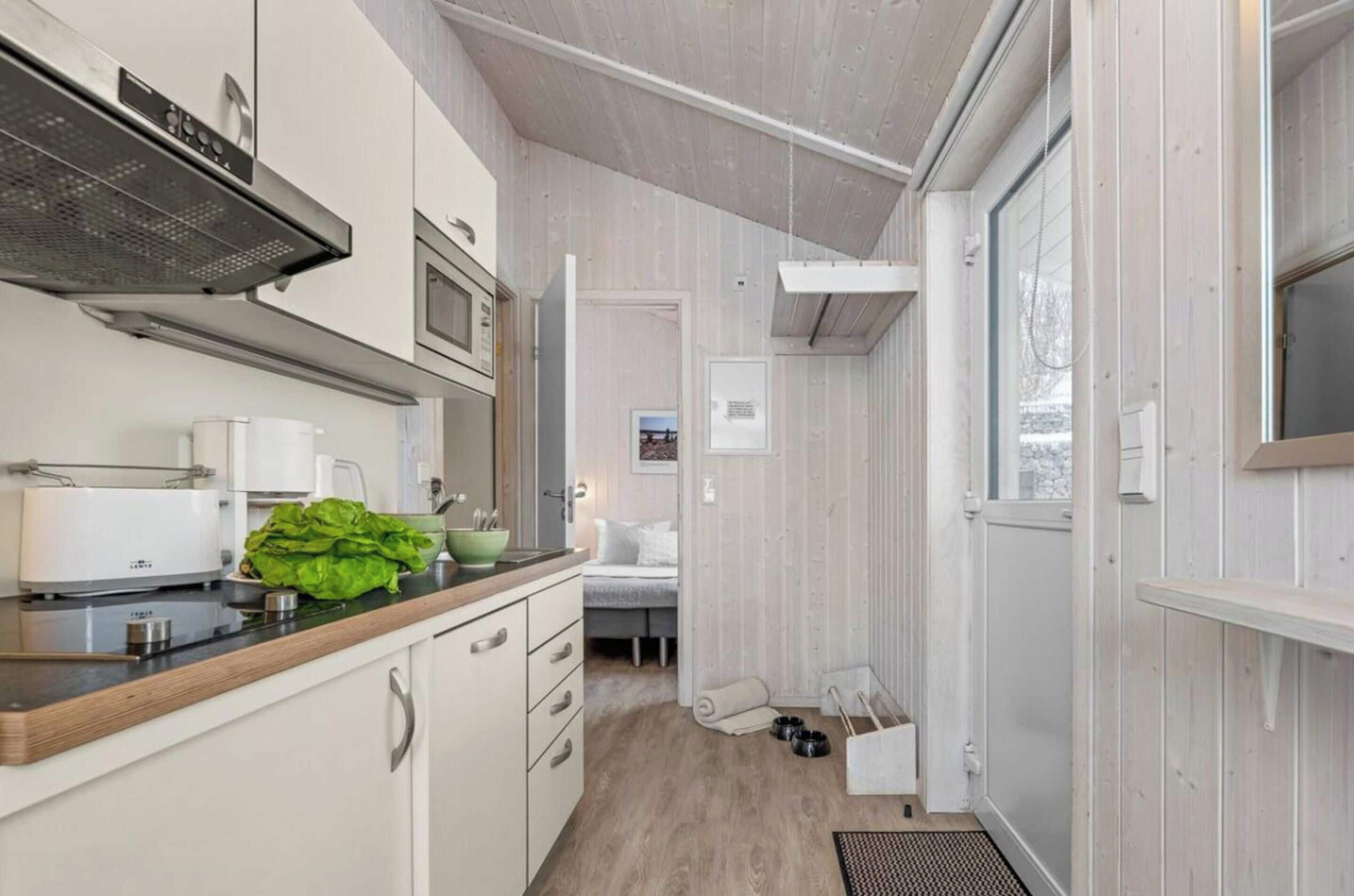 Et kompakt køkken med moderne hårde hvidevarer, et spiseområde med bord og stole, og et lille soveområde med seng og hundeseng.