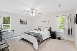 A bedroom with a bed, a desk, a chair, a ceiling fan, and a window with curtains.