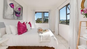 Ein Schlafzimmer mit einem Bett, Kissen, einem Gemälde, einem Fenster und Blick ins Freie.