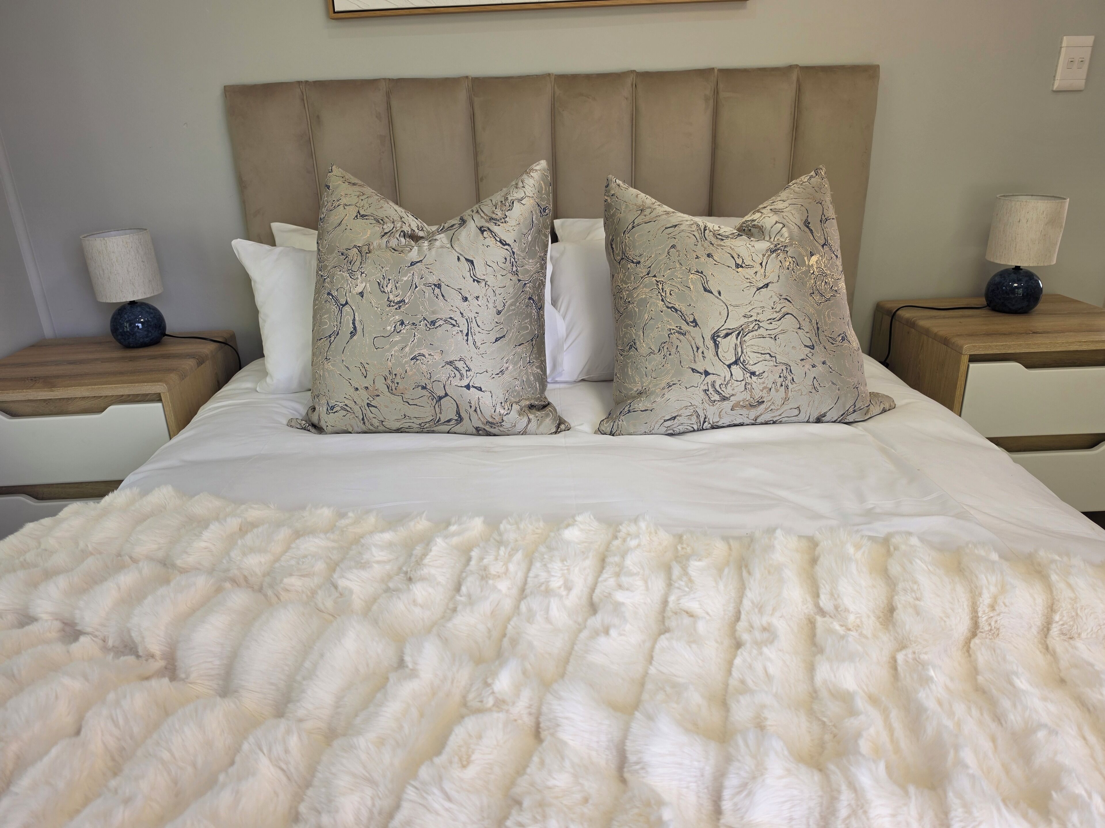 Una cama con edredón blanco y dos almohadones decorativos, flanqueada por mesitas de noche de madera con lámparas.