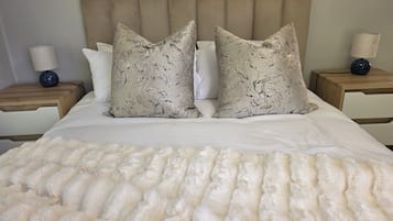 Una cama con edredón blanco y dos almohadones decorativos, flanqueada por mesitas de noche de madera con lámparas.