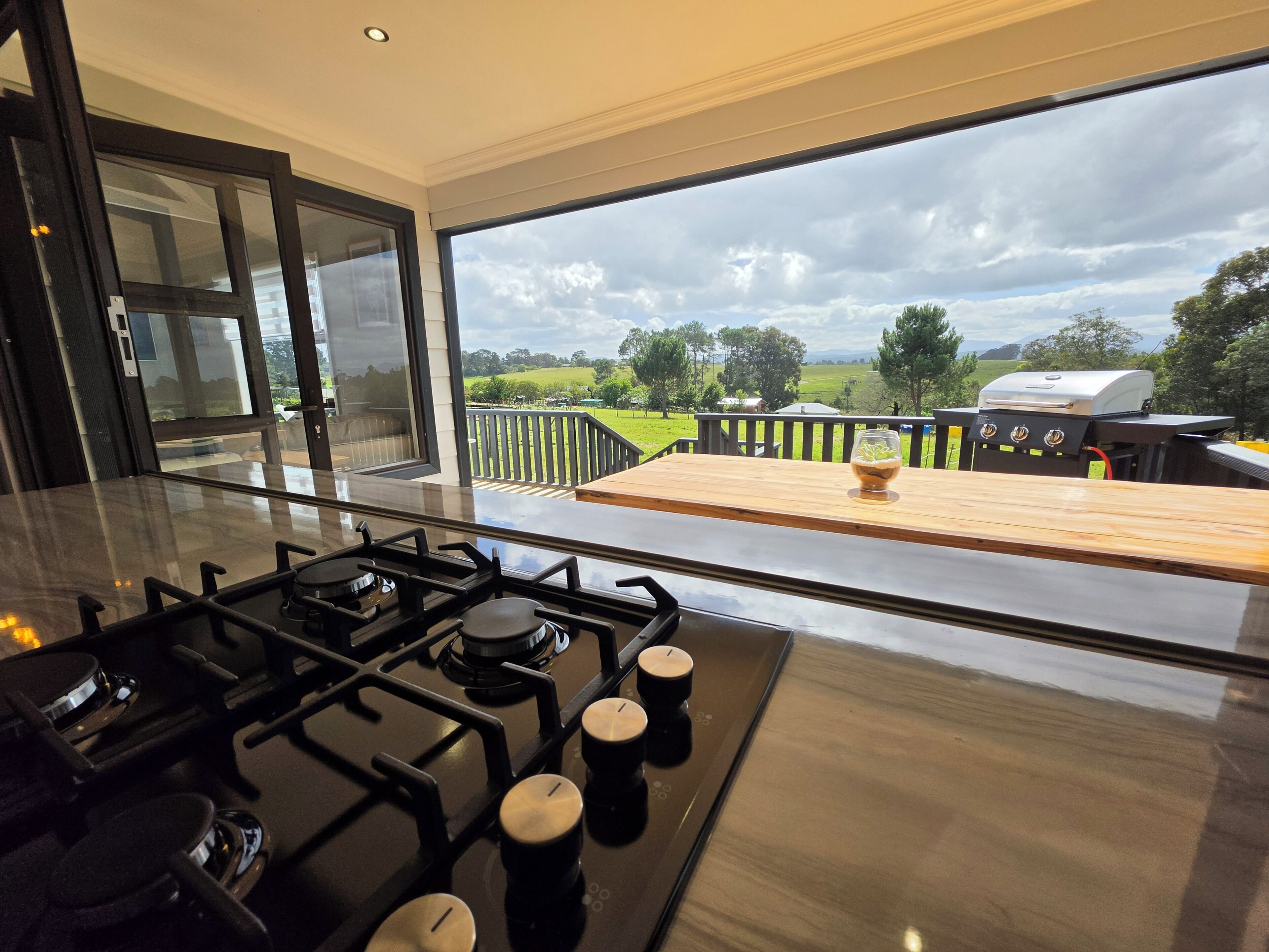 Cocina moderna con estufa a gas, mesa de madera y vista a una parrilla y al paisaje.