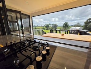 A modern kitchen with a gas stove, a wooden table, and a view of a barbecue grill and landscape.