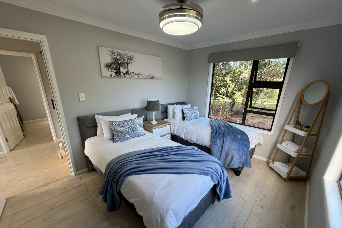 Quarto com duas camas, uma mesa de cabeceira com abajur, um quadro na parede e uma janela com vista para árvores.