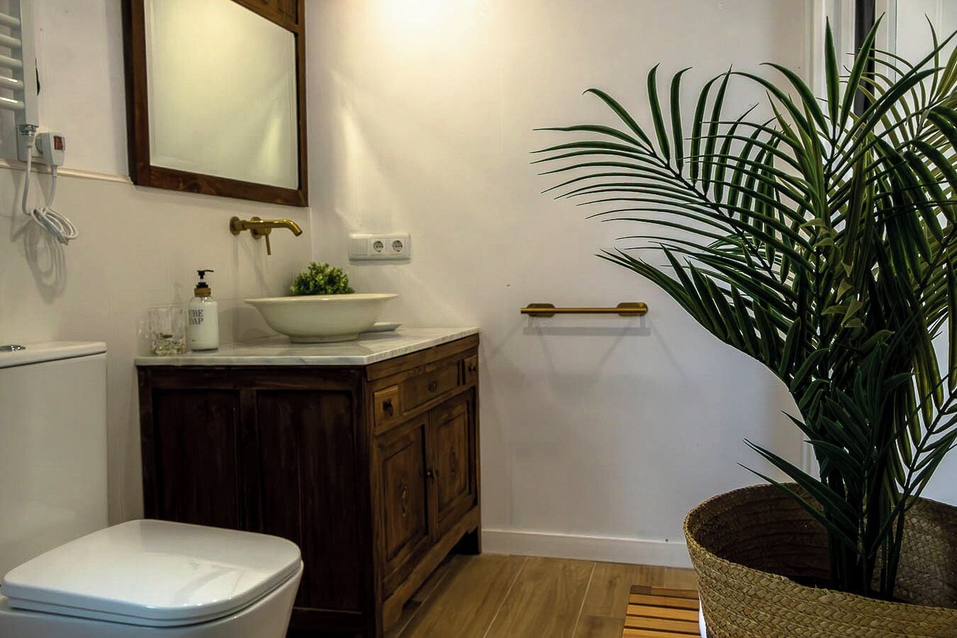 A bathroom with a toilet, a sink with a bowl, a mirror, a plant, and a wooden cabinet.