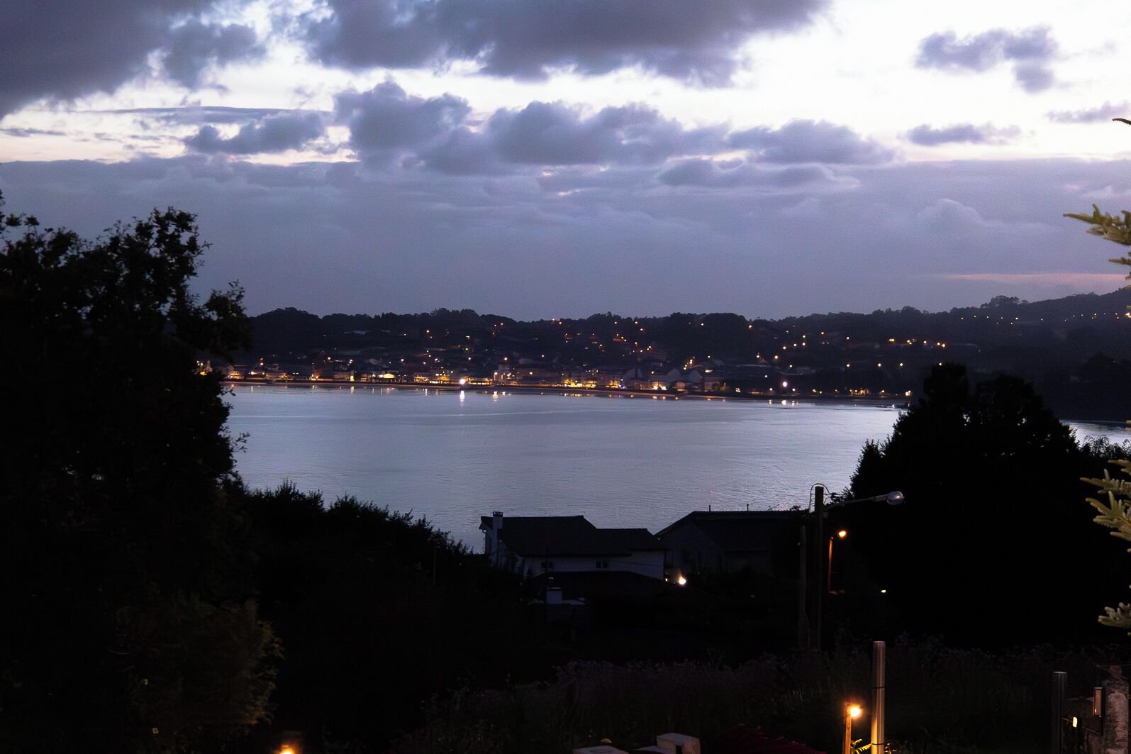 A coastal view with a body of water, a distant shoreline with lights, and a cloudy sky.