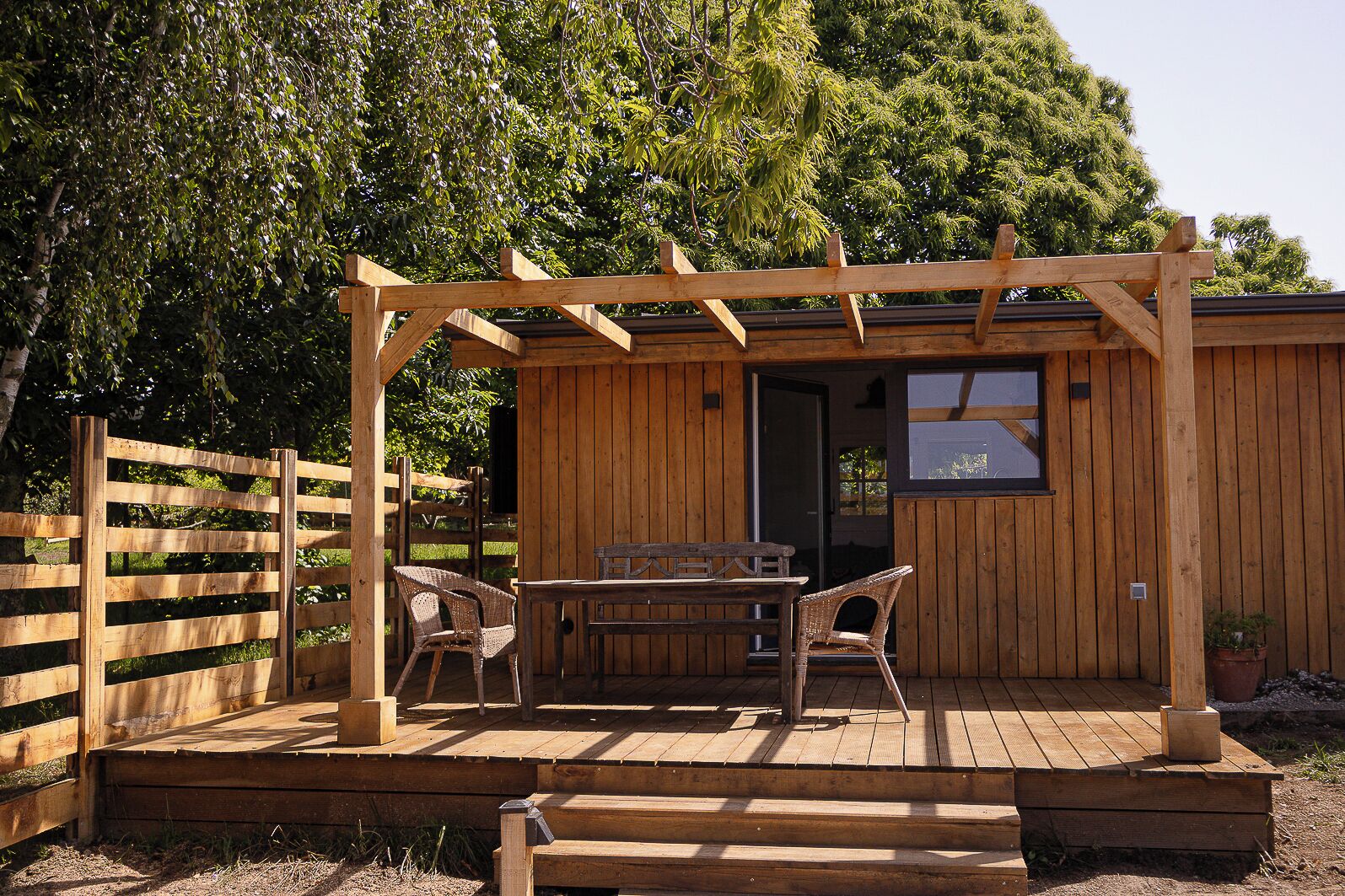 Una terraza de madera con una mesa y sillas, un pérgola de madera y una construcción de madera con un ventanal.