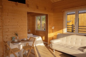 Interior de una cabaña de madera con comedor, un sofá y ventana con vistas a follaje.