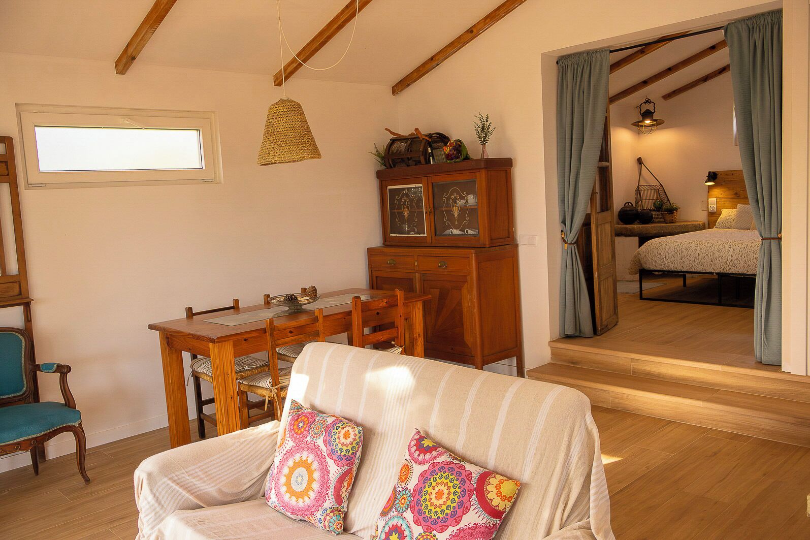 A cozy living room with a dining table, a sofa with colorful cushions, a wooden cabinet, and a view into a bedroom with a bed and curtains.