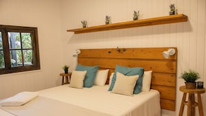 A bedroom with a wooden headboard, a bed with white bedding, multiple pillows, a bedside table with a lamp, a window, and a shelf with plants. - Raíces da costa Ártabra (A Coruña)