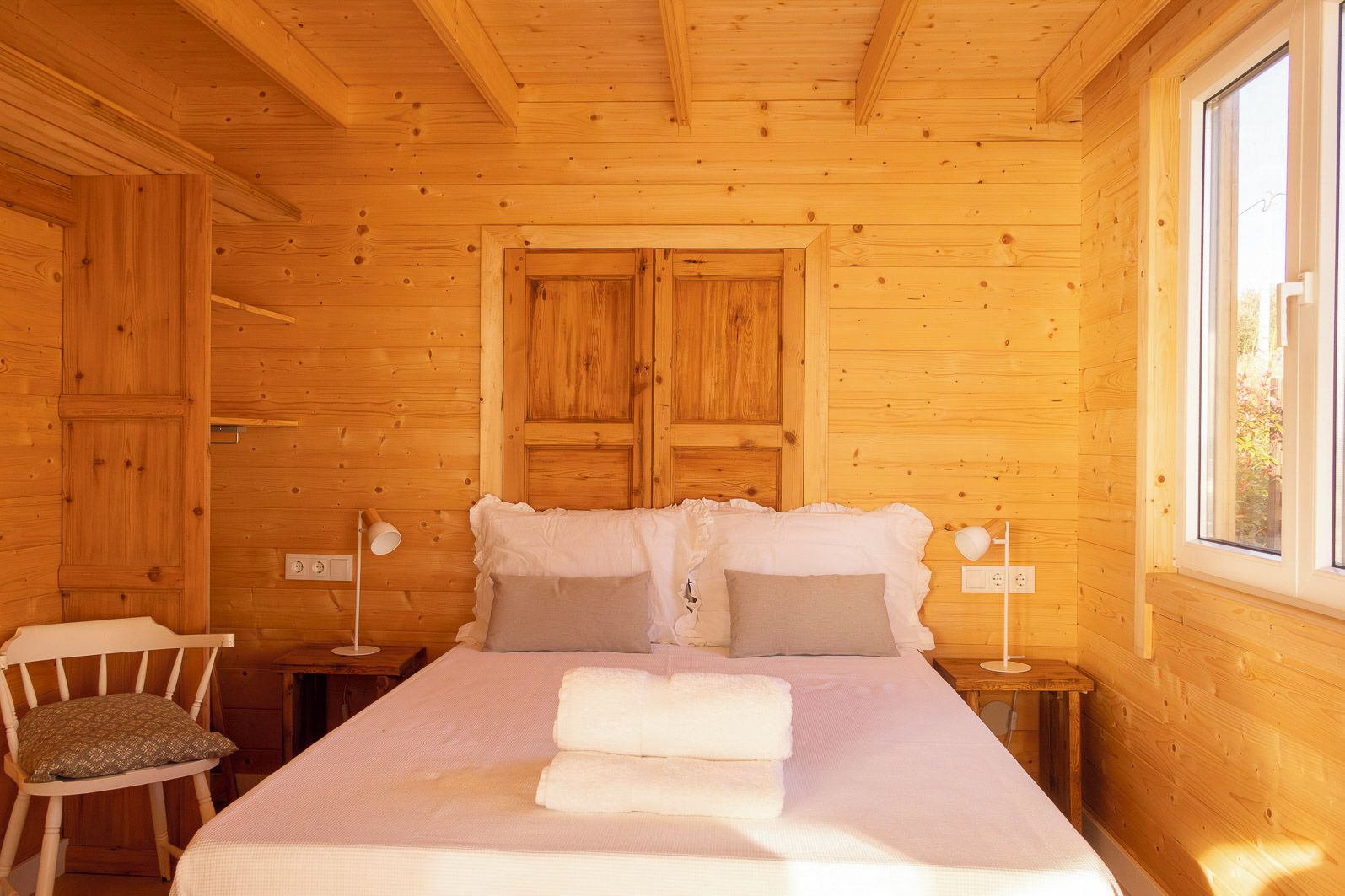 Ett sovrum i en stuga med träpanel, inrett med en säng, två sänglampor, en stol och ett fönster.