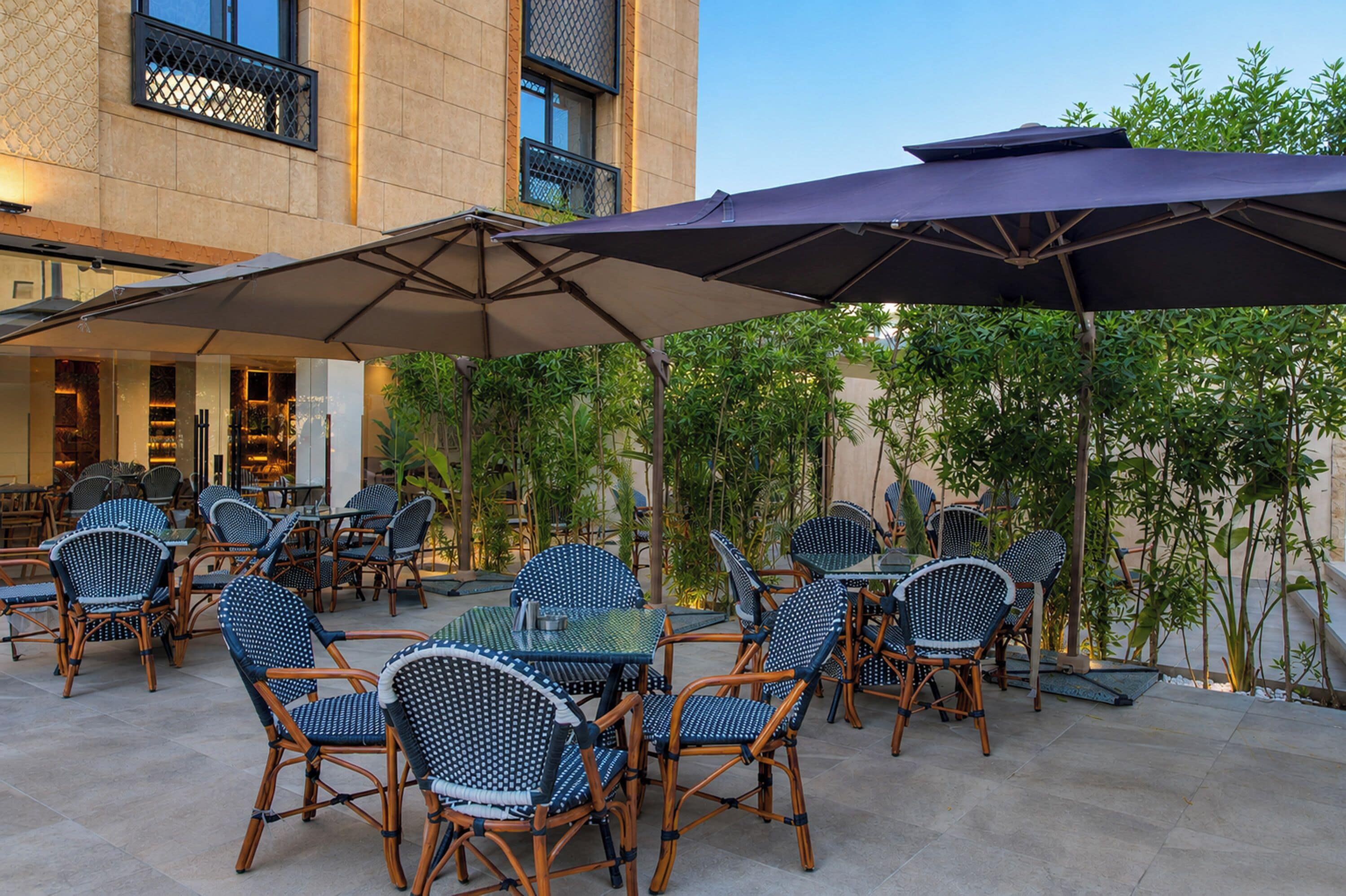 An outdoor seating area with wicker chairs and tables, shaded by large umbrellas, in front of a building with a tiled facade and a balcony.