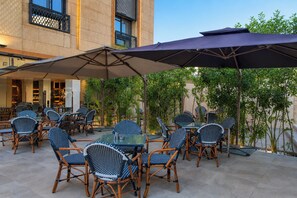 An outdoor seating area with wicker chairs and tables, shaded by large umbrellas, in front of a building with a tiled facade and a balcony. - Touring Pyramids Hotel (Giza)
