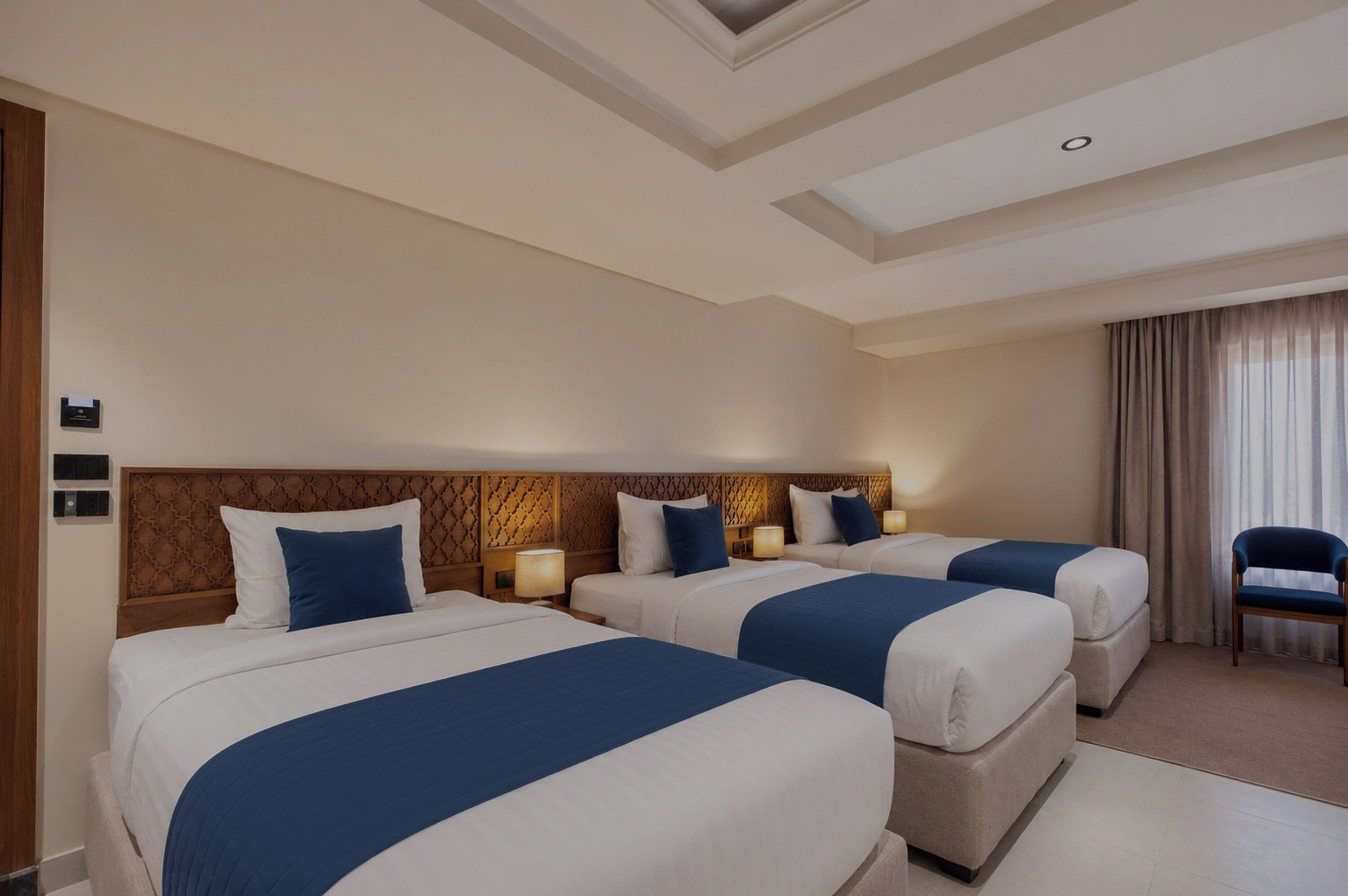 Habitación de hotel con dos camas, cabecero de madera, mesita de noche con lámpara y una silla.