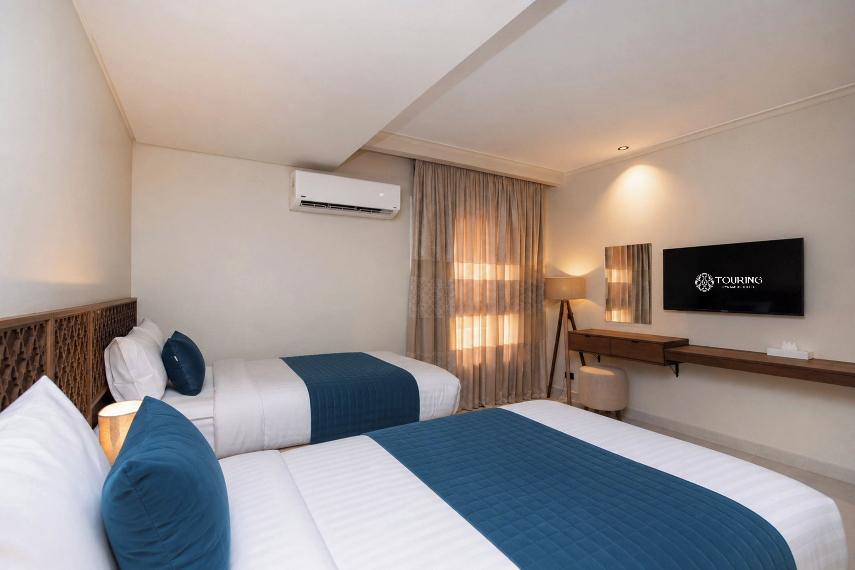 A hotel room with two beds, a TV, a desk, and an air conditioning unit.