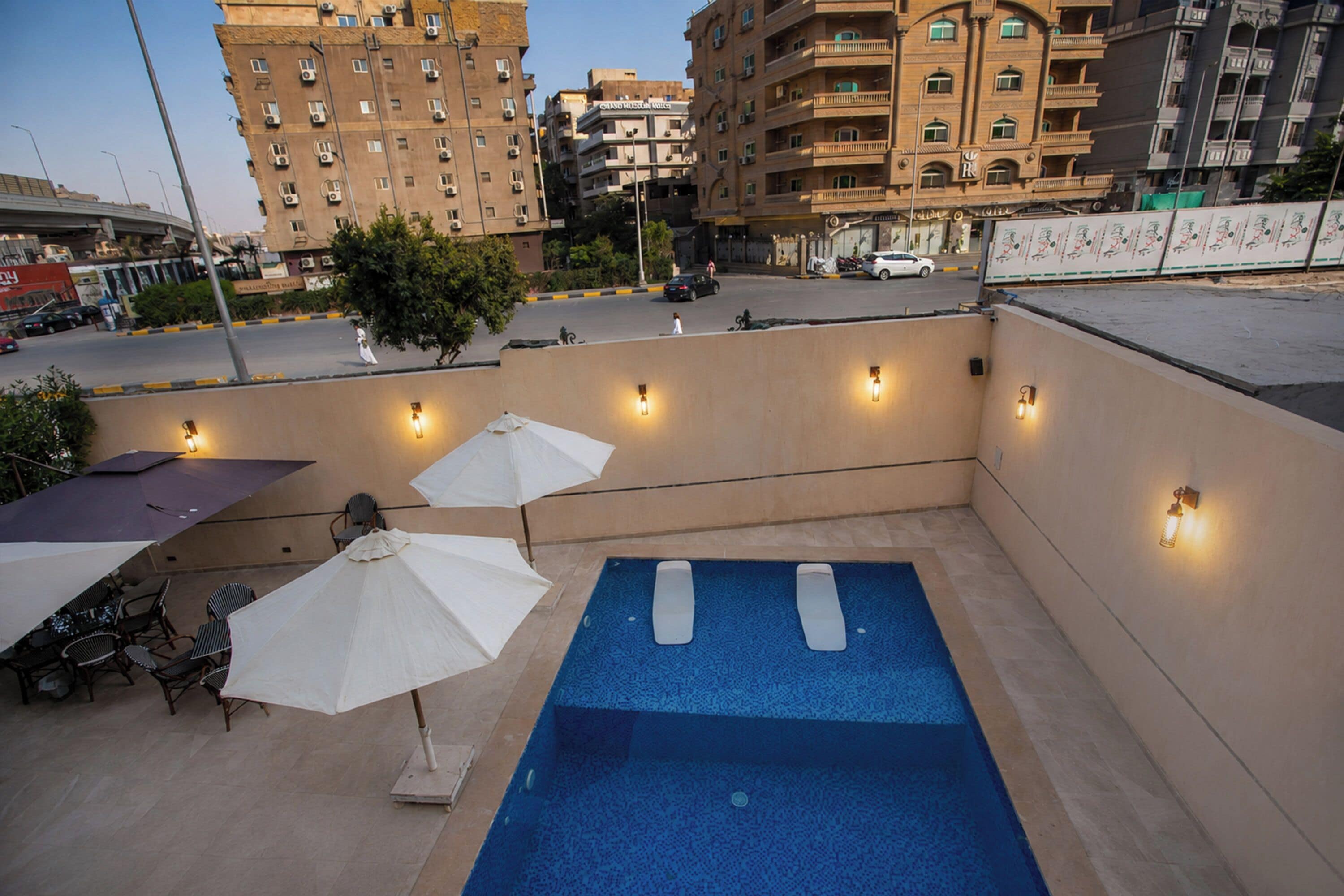 Un área de piscina en la azotea con sillones de descanso y sombrillas, rodeada de edificios.