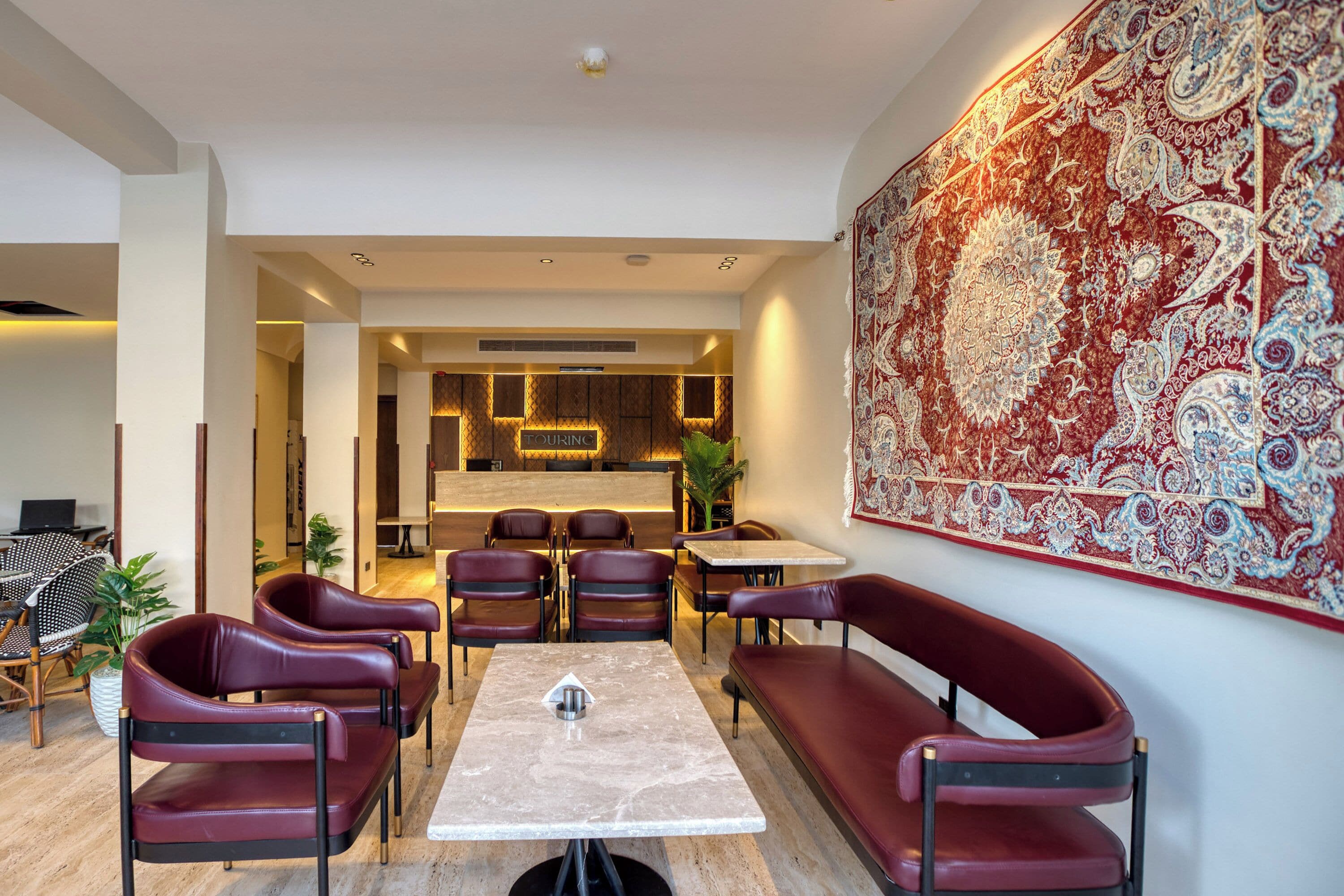 Un interior moderno con una zona de comedor que cuenta con una mesa de mármol y sillas de cuero, un tapiz grande y ornamentado en la pared, y un mostrador de recepción al fondo.