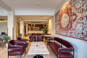 Un interior moderno con una zona de comedor que cuenta con una mesa de mármol y sillas de cuero, un tapiz grande y ornamentado en la pared, y un mostrador de recepción al fondo.