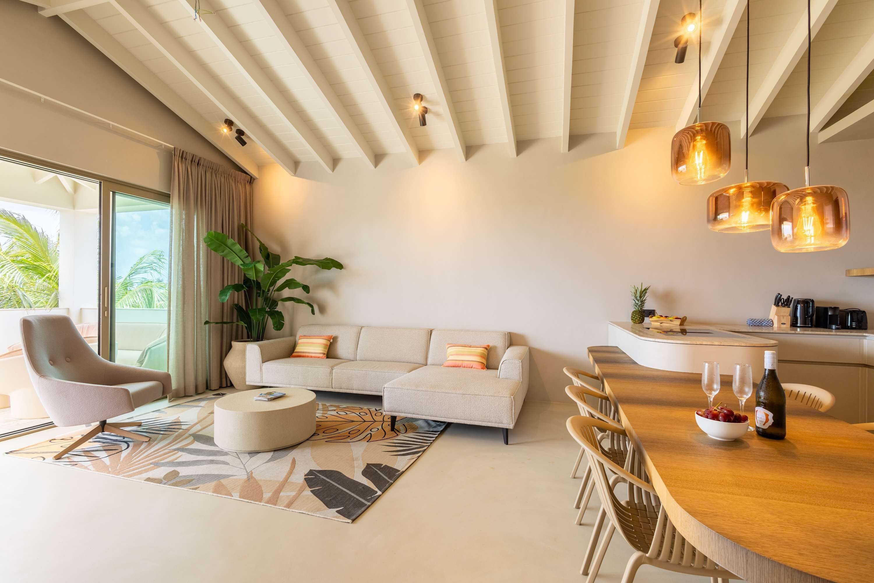 A modern living room with a sofa, a coffee table, and a dining area with chairs. There is a large window with curtains, a plant, and a kitchen area with a counter and appliances.