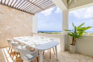 A terrace with a table set for dining, white chairs, a potted plant, and a view of the sea. - Azzurro at The Reef Ocean View Penthouse (Sint Michiel)
