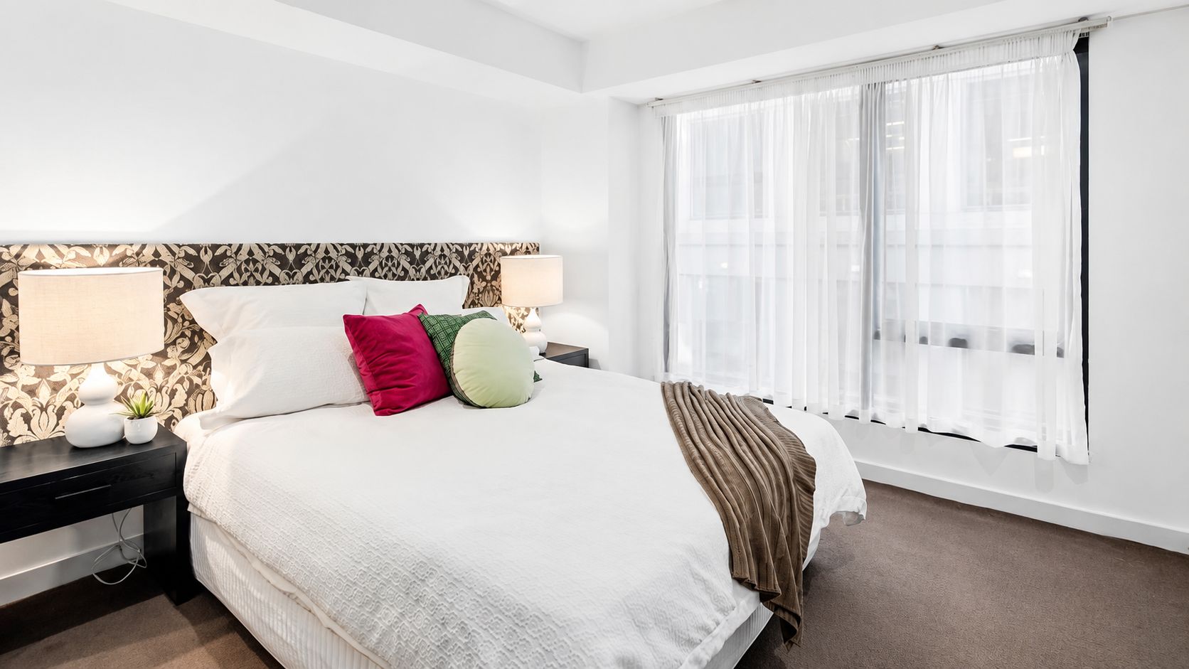 A hotel room with a large bed, two bedside tables, a window with sheer curtains, and a patterned wallpaper on the headboard.