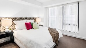 A hotel room with a large bed, two bedside tables, a window with sheer curtains, and a patterned wallpaper on the headboard. - Spacious 70m² St Kilda Rd Stay Near Alfred Hospital with Gym,Parking & Workspace (Melbourne)