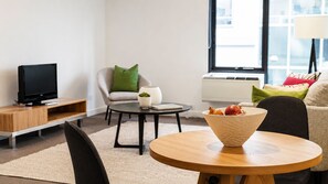 A modern living room with a flat-screen TV, a round wooden table, a grey armchair, a small round coffee table, and a sofa with cushions. - Spacious 70m² St Kilda Rd Stay Near Alfred Hospital with Gym,Parking & Workspace (Melbourne)