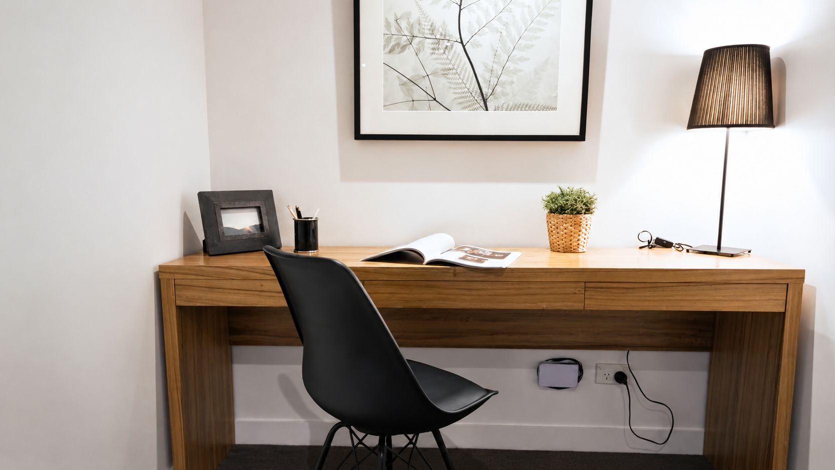 Un bureau en bois, une chaise noire, un tableau encadré, une petite plante et une lampe.