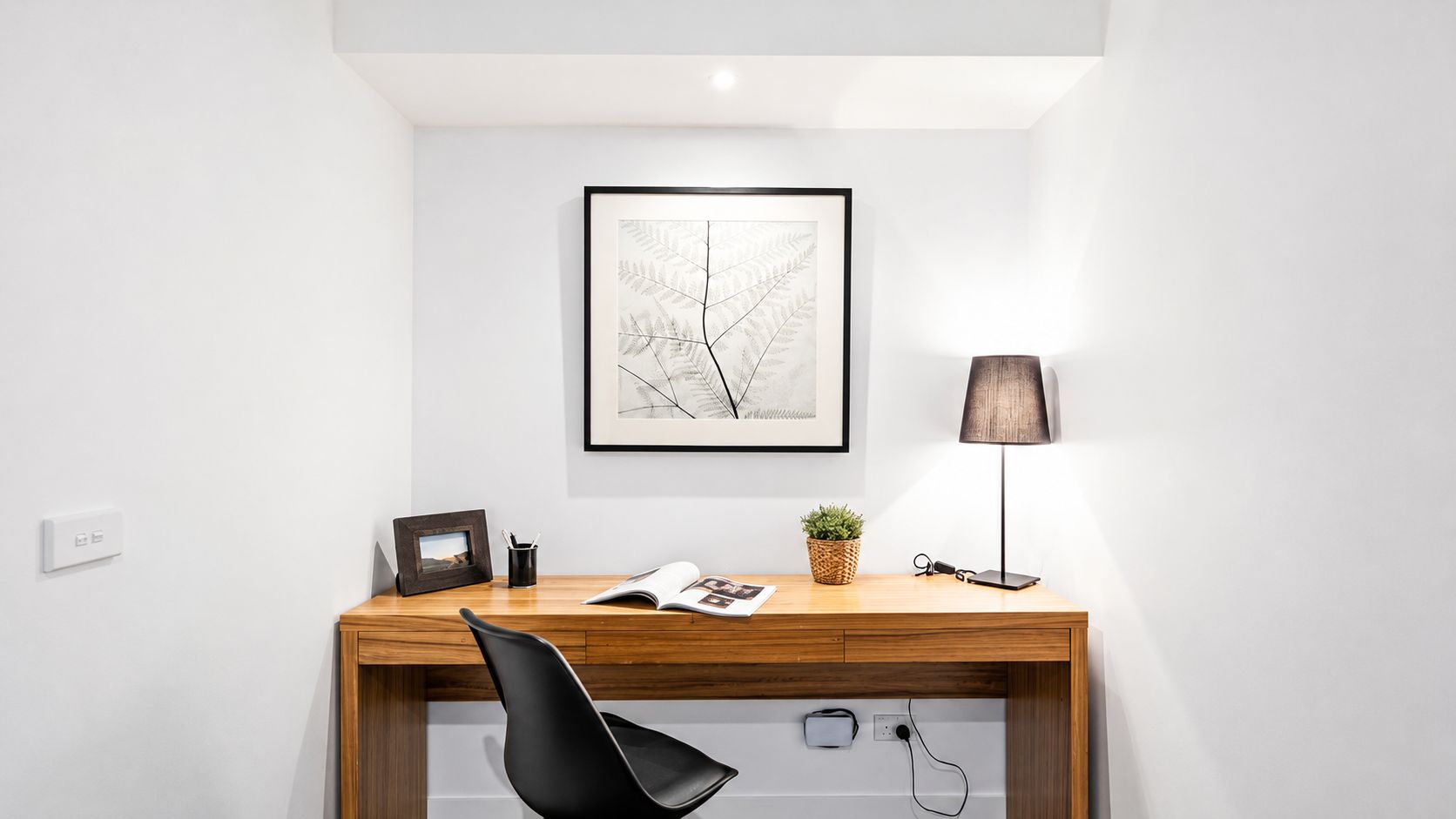 Un espace de bureau moderne avec un bureau en bois, une chaise noire, un tableau encadré, une lampe et une plante en pot.