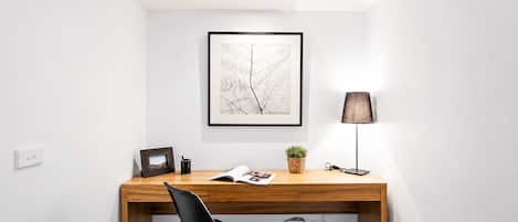 A modern office space with a wooden desk, a black chair, a framed picture, a lamp, and a potted plant.