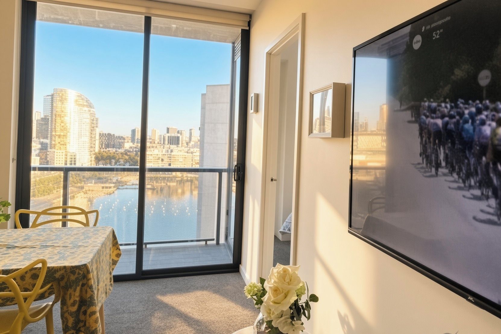 A room with a large window, a dining table with chairs, a flower vase, and a television screen displaying a crowd.