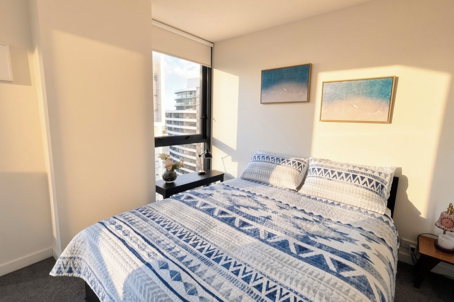 A bedroom with a bed, a nightstand, a window with a view of buildings, and two wall-mounted pictures.