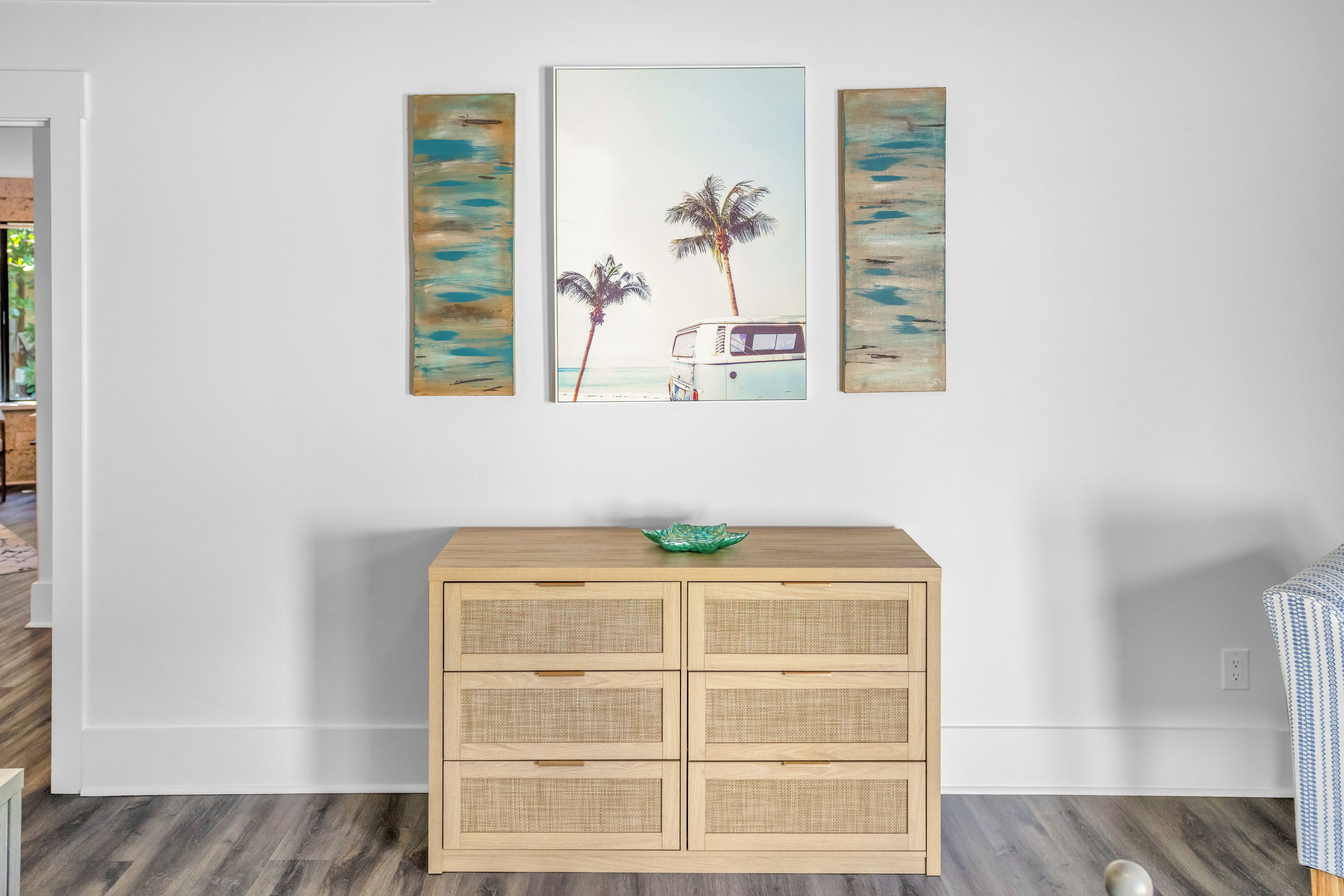 Una habitación con un armario de madera, un cuadro colgado en la pared que muestra una playa y un cuenco verde sobre el armario.