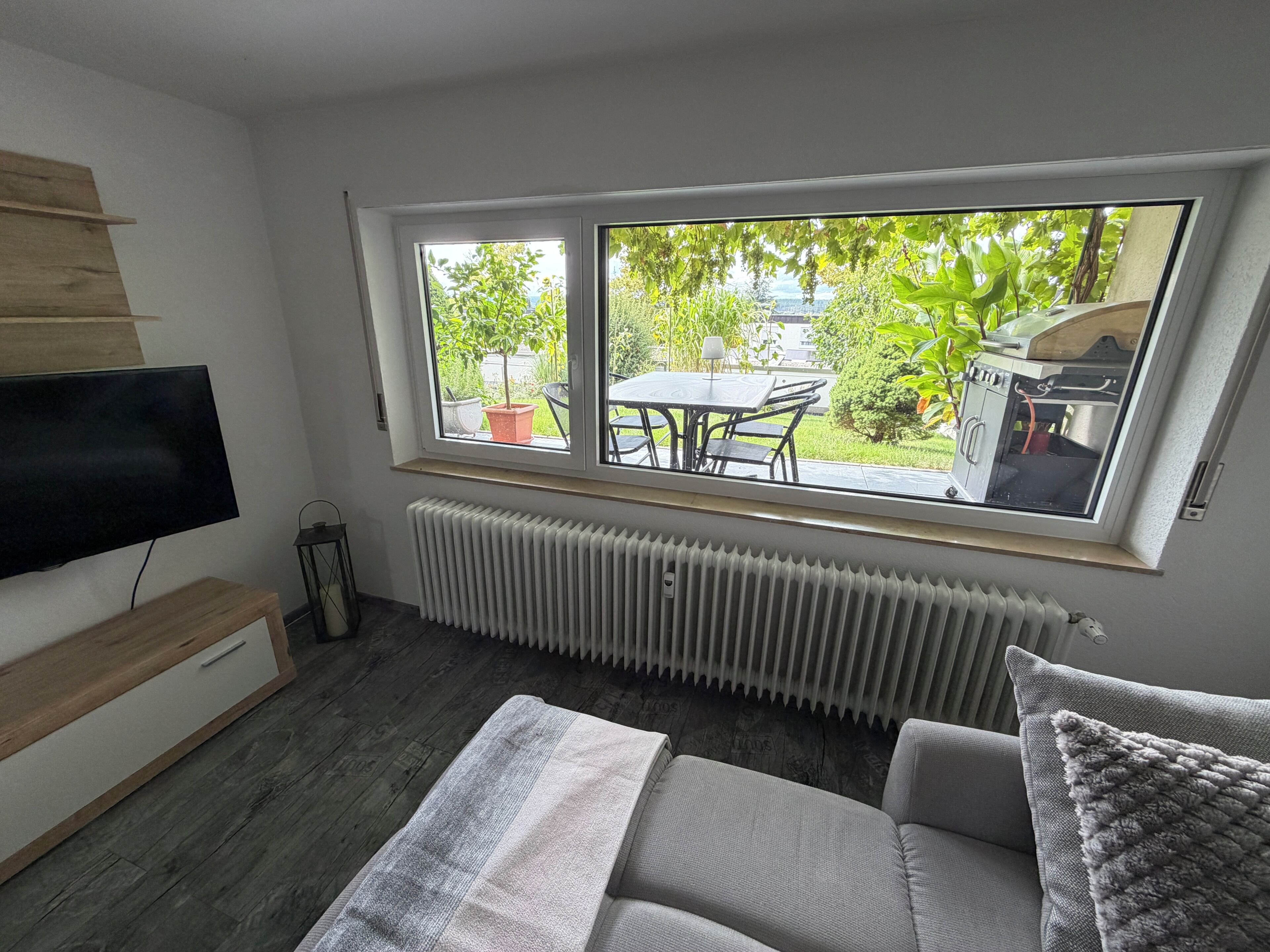 Ein Zimmer mit großem Fenster, Flachbildfernseher, Heizkörper und einer Couch.