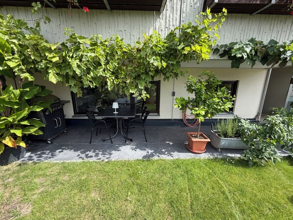 Un patio al aire libre con una mesa y sillas, rodeado de follaje y una pared blanca.